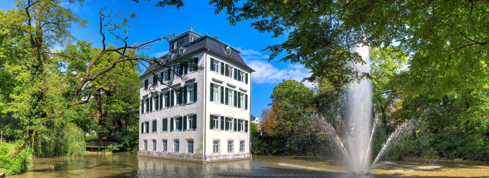 Im Blickfeld ist das Holzhaussenschlössen im See mit großen Springbrunnen. Links und Rechts grüne Bäume.