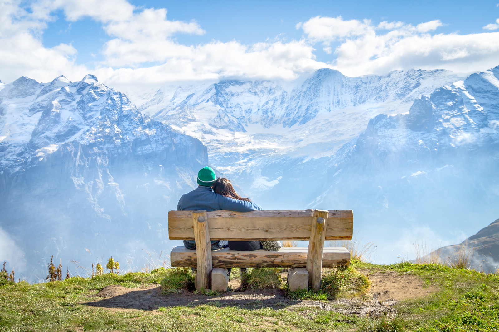 Ein Paar das auf einer Holzbank sitzt und ein tollen Blick auf die Schnee bedeckten Alpen hat.