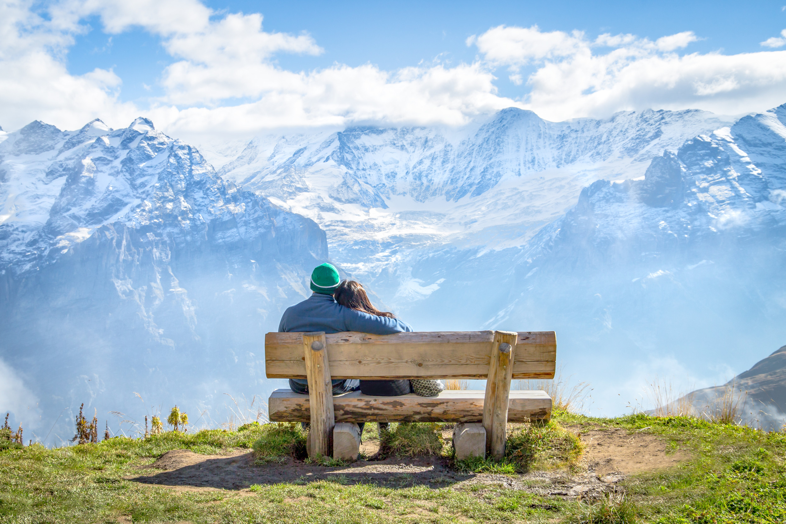 Ein Paar das auf einer Holzbank sitzt und ein tollen Blick auf die Schnee bedeckten Alpen hat.
