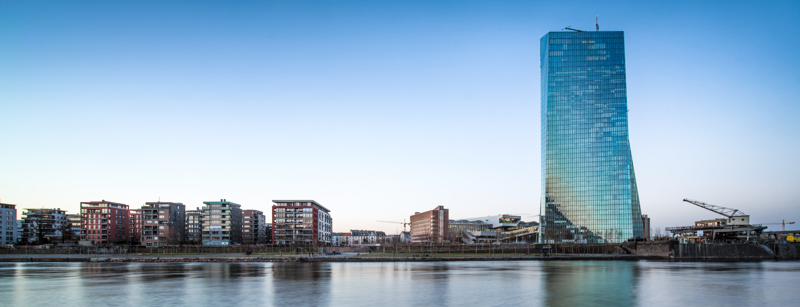 Im Vordergrund der Fluss Main. Im Hintergrund das neue Gebäude der EZB und daneben kleine Wohnhäuser bei blauen Himmel