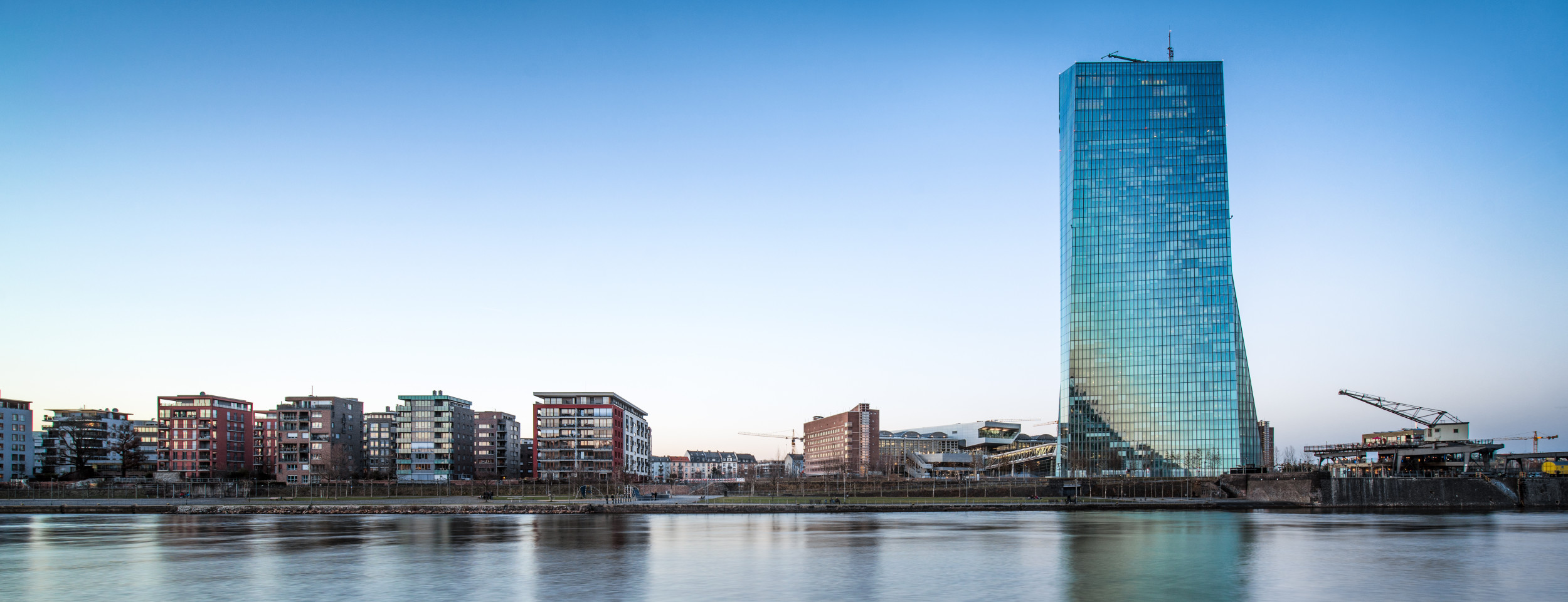 Im Vordergrund der Fluss Main. Im Hintergrund das neue Gebäude der EZB und daneben kleine Wohnhäuser bei blauen Himmel